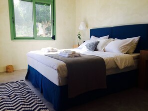 Una cama bien tendida con ropa de cama blanca y azul, un tapete a rayas, una ventana con marco verde y una lámpara de noche.
