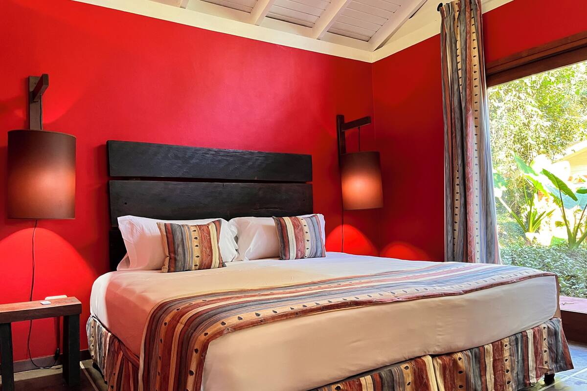 A bedroom with a red wall, a wooden headboard, a bed with striped bedding, two bedside lamps, and a window with curtains.