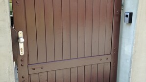 Una verja metálica marrón con una cerradura y un ojo de cerradura, unida a una pared beige con una pequeña caja eléctrica cerca.