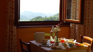 Une table à manger dressée avec des aliments pour le petit-déjeuner et un vase de fleurs, le tout placé devant une fenêtre offrant une vue sur les montagnes.