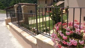 Un bâtiment en pierre, doté d’un toit en tuiles, entouré d’une clôture en métal et de plantes en fleurs.