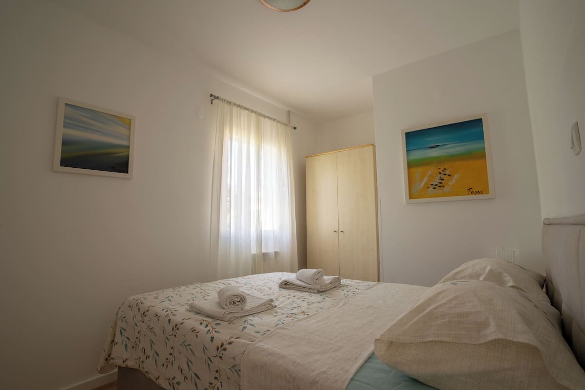 A bedroom with a bed, a window with curtains, and two paintings on the walls.