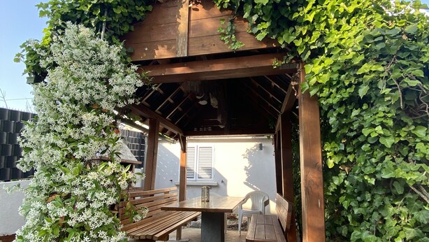 Una pergola in legno con una panca e un tavolo, circondata da vegetazione rigogliosa e fiori bianchi.