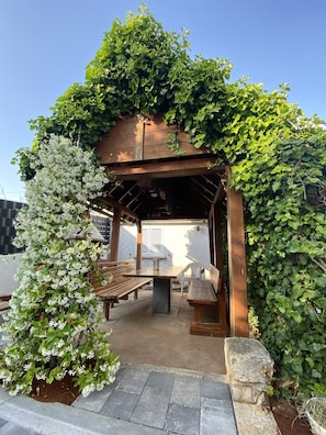 A wooden pergola with a bench and table, surrounded by lush greenery and white flowers. - Apartments & Rooms Petra (Zadar)