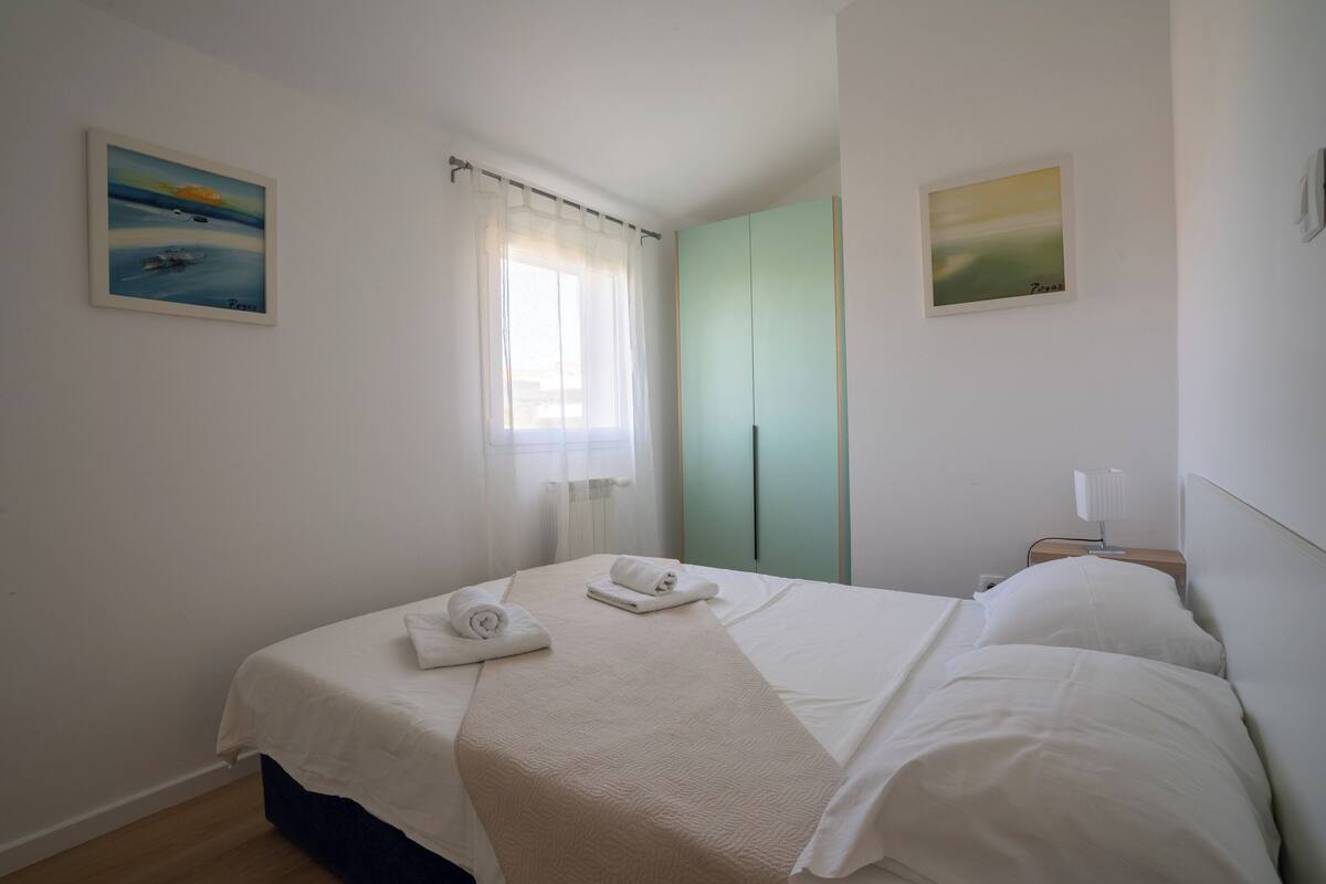 A neatly made bed with white linens and towels, a window with sheer curtains, and a green wardrobe.
