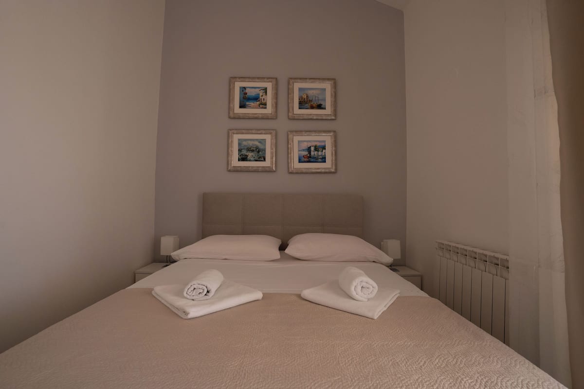 A neatly made bed with two pillows, a headboard, and two rolled towels on the bed. There are four framed pictures on the wall above the bed.