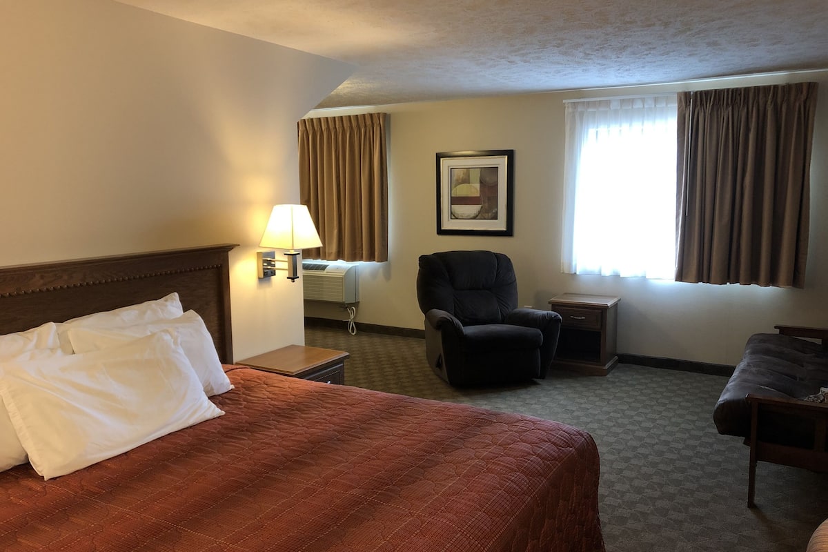A hotel room with a large bed, a chair, a desk, and a window with curtains.