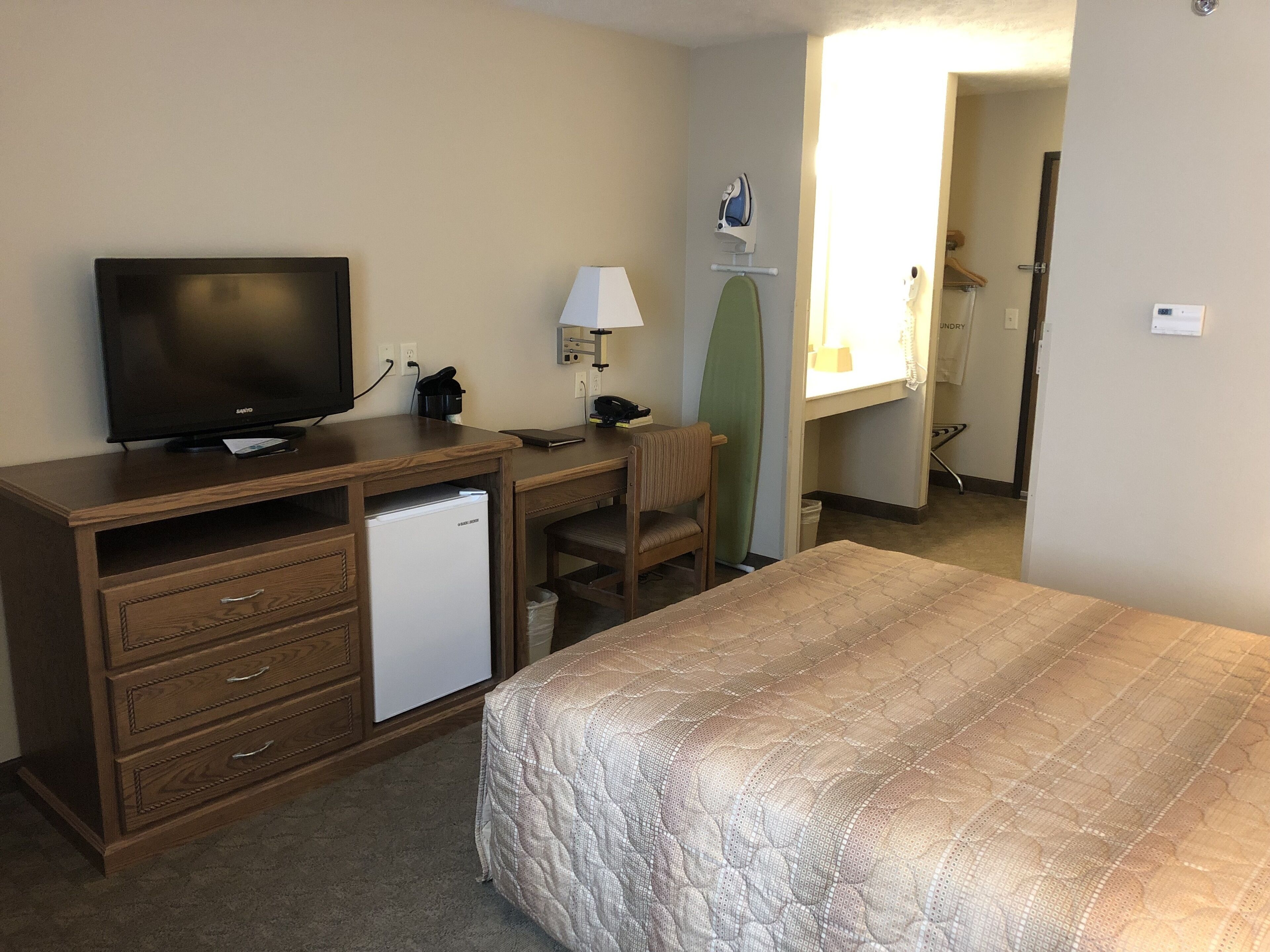 A hotel room with a bed, a desk with a television, a mini-fridge, a chair, and a surfboard.