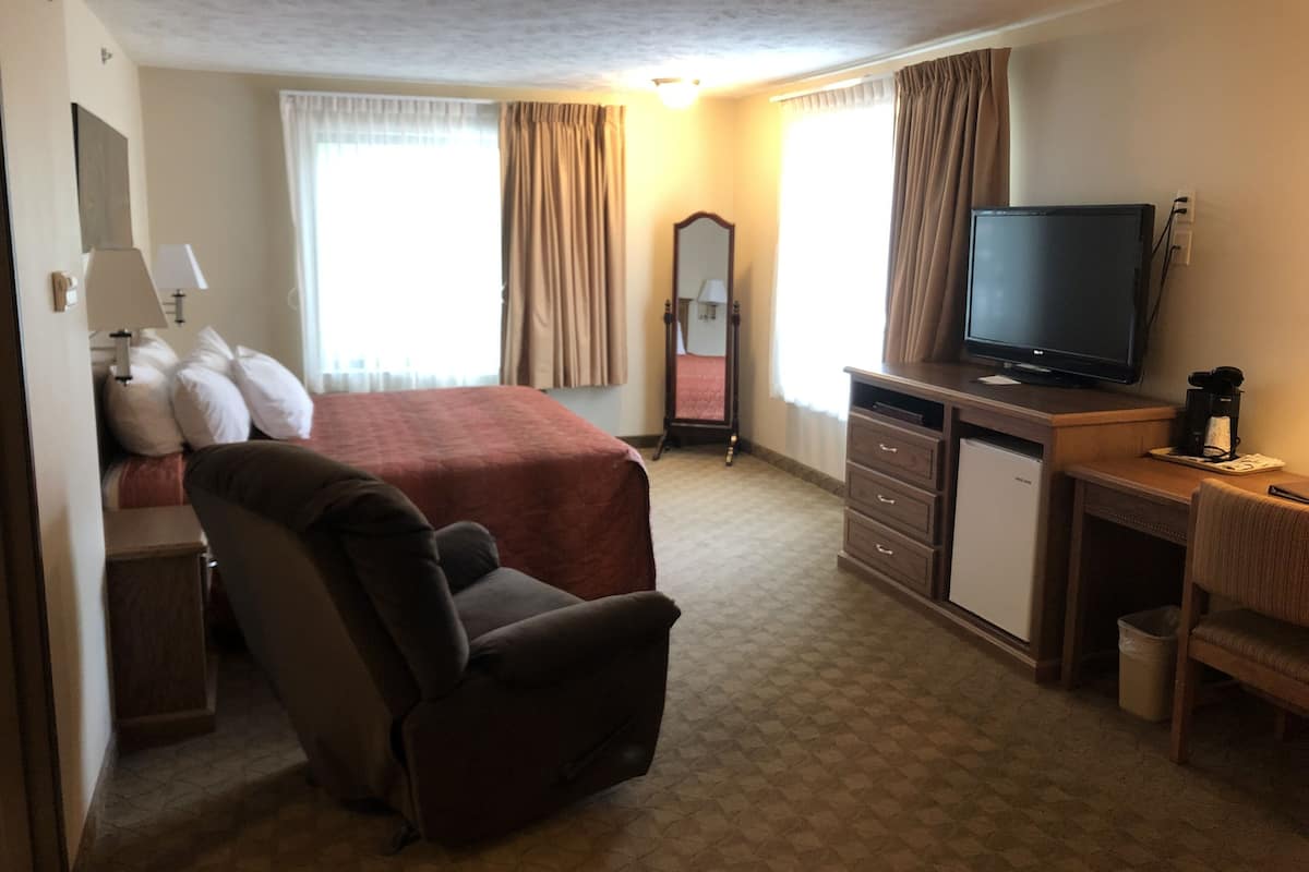 A hotel room with a bed, a chair, a desk with a television, a mirror, and a dresser.