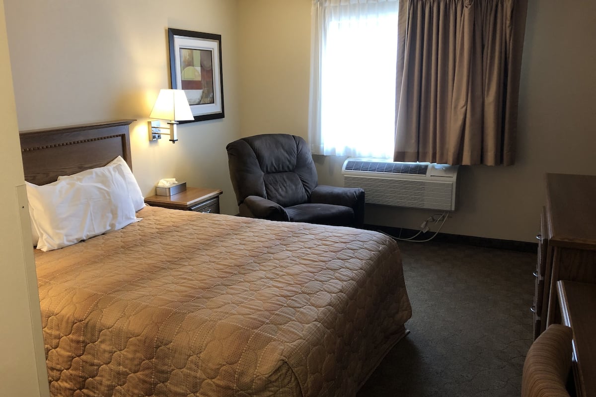 A hotel room with a large bed, a chair, a desk, an air conditioning unit, and a window with curtains.