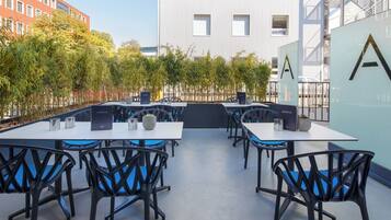 Un área de estar al aire libre con mesas y sillas, rodeada de follaje y un edificio moderno al fondo.