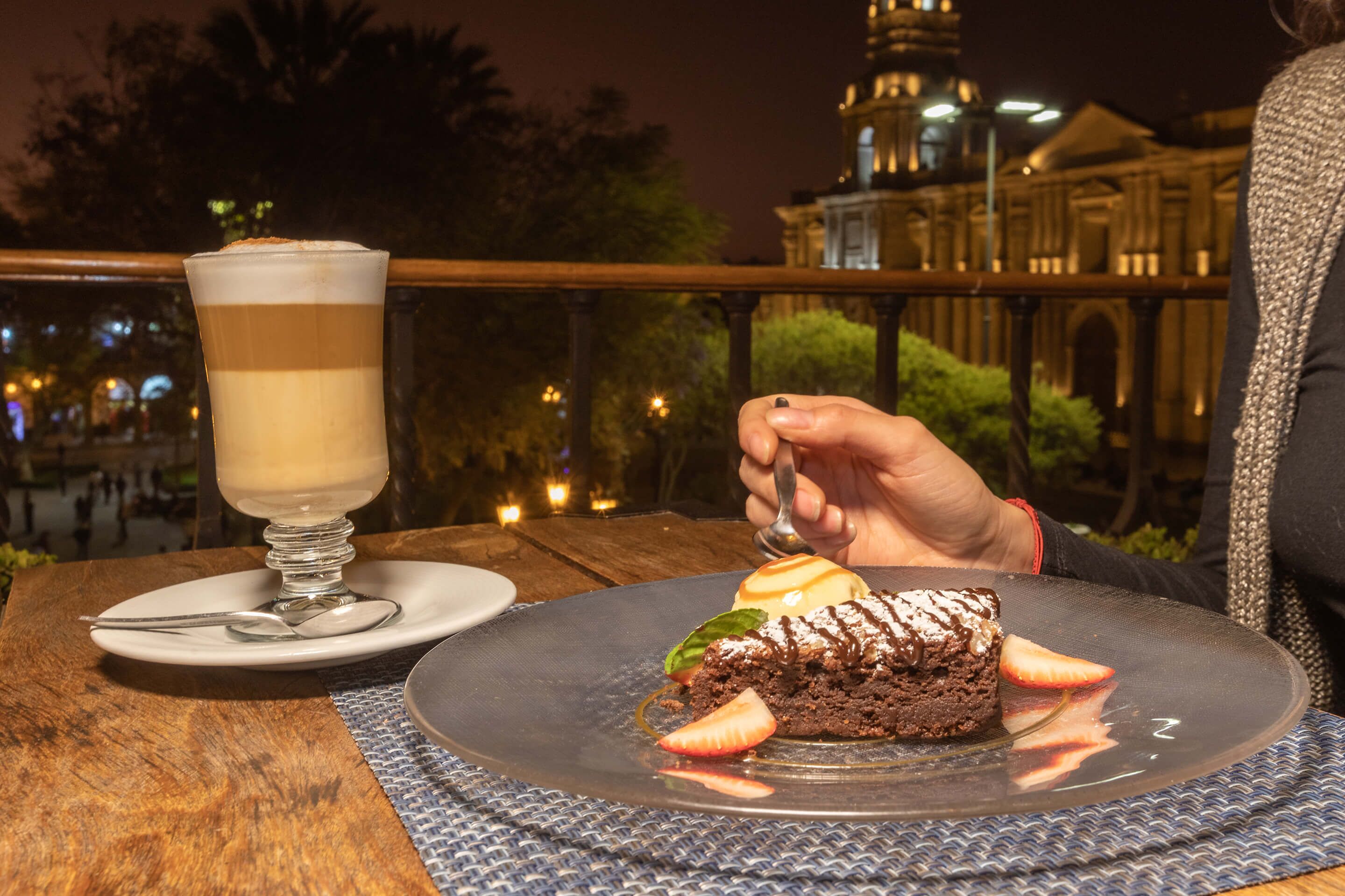 Un plato de postre con un pastel de chocolate y fresas, un vaso de una bebida en capas, y una persona sosteniendo una cuchara.