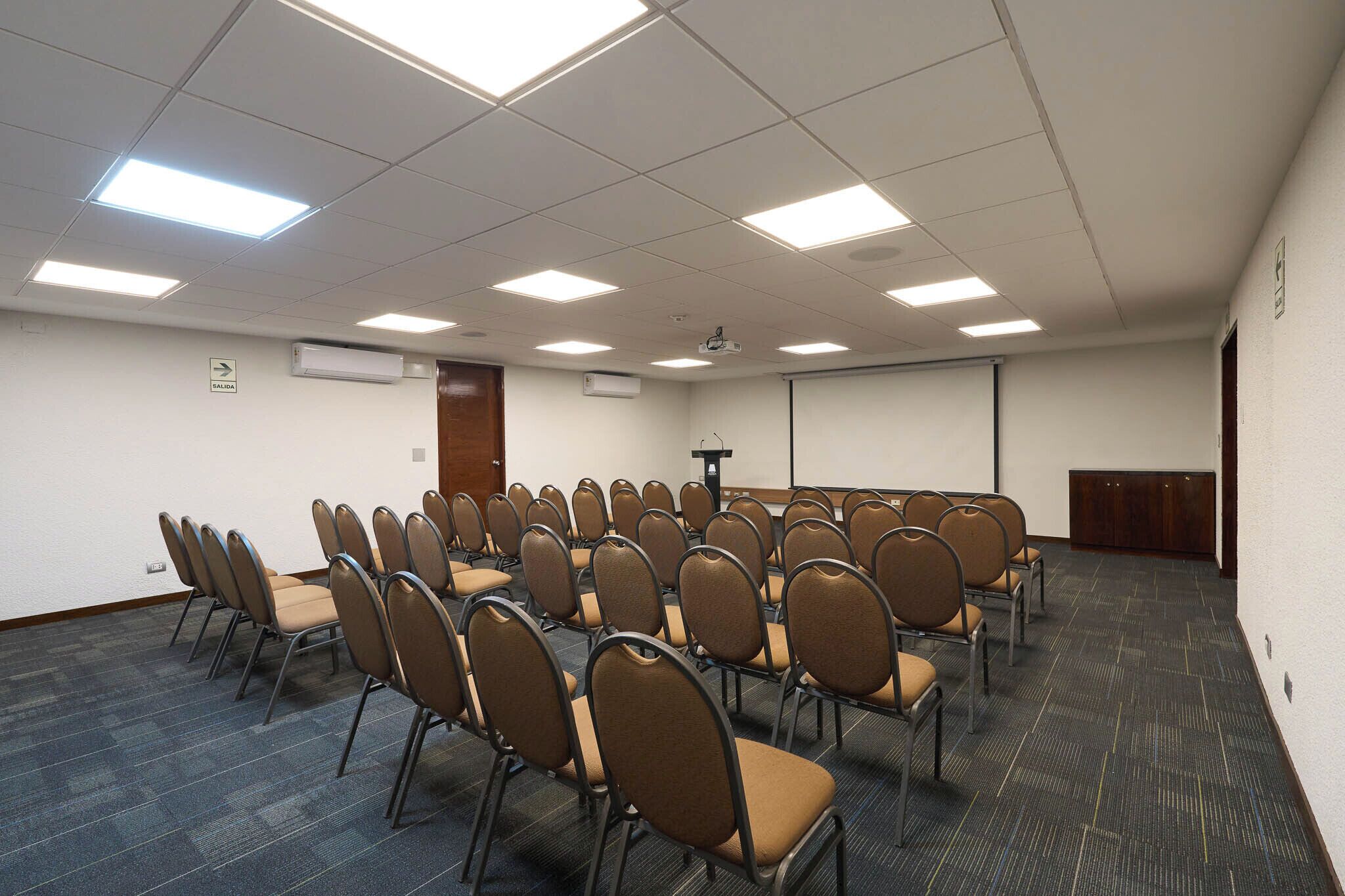 Un salón de conferencias con filas de sillas, luces en el techo y un equipo de aire acondicionado.