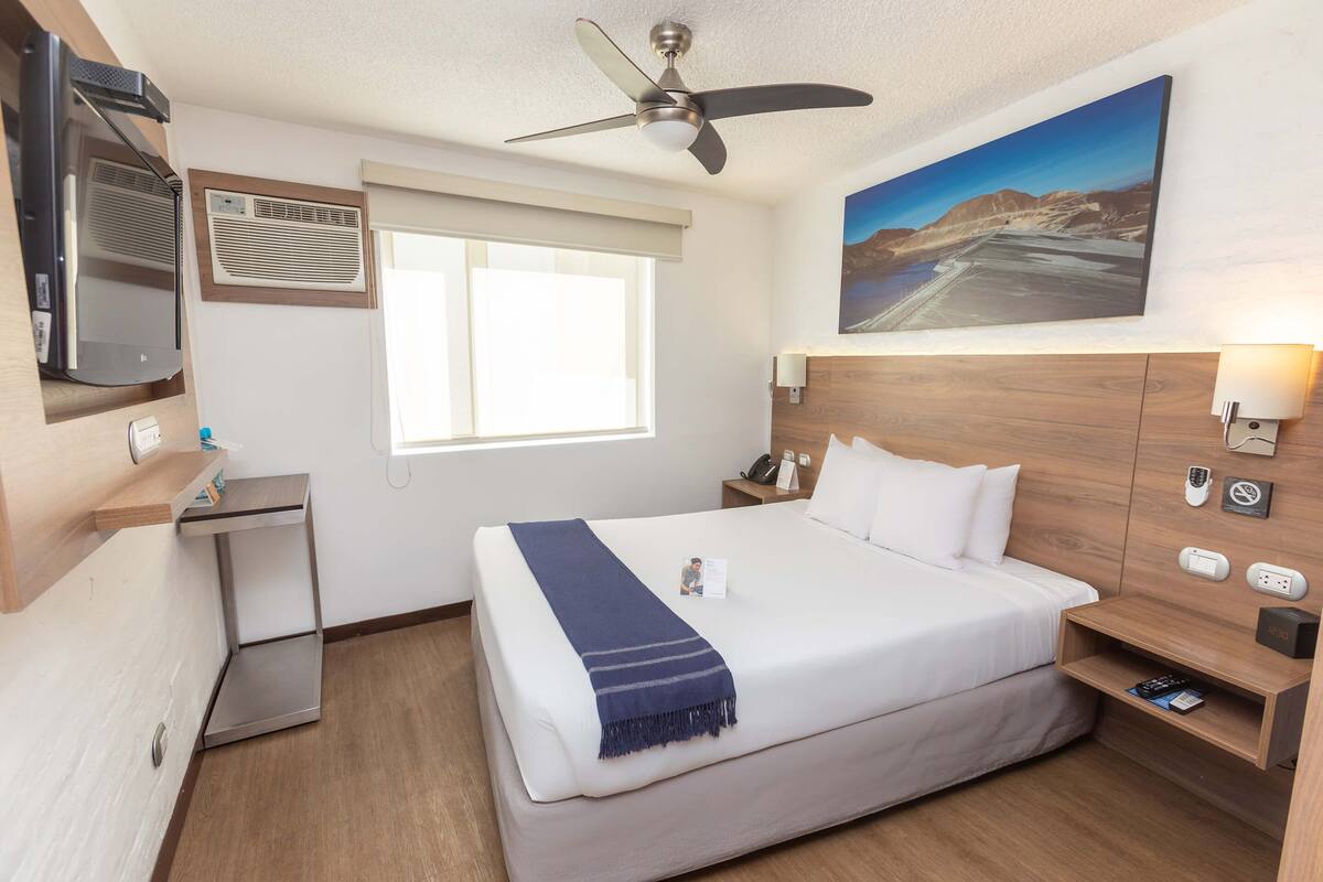 Ein Hotelzimmer mit Bett, Schreibtisch, Fernseher, Deckenventilator, einem Gemälde und einem Nachttisch mit Lampe.