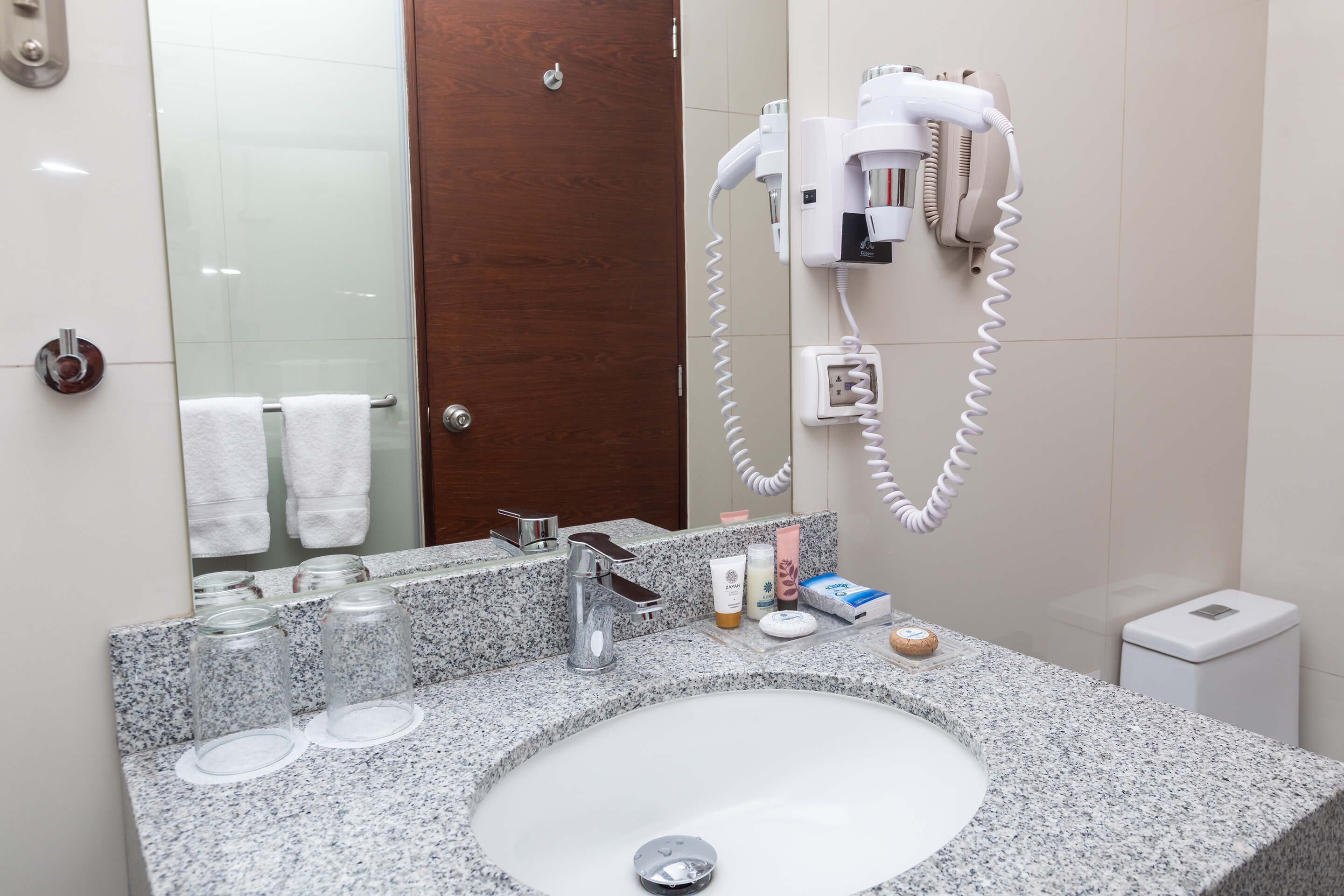 A bathroom with a granite countertop, a sink, a mirror, a hairdryer, and a small trash bin.