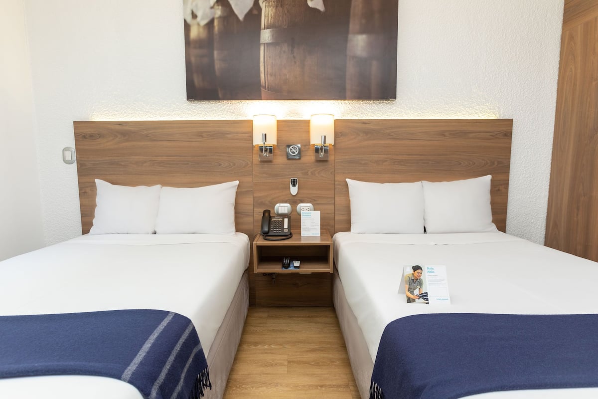 Ein Hotelzimmer mit zwei Einzelbetten, einem Holzkopfteil, Nachttischen mit Lampen, einem Telefon und einem kleinen Regal.