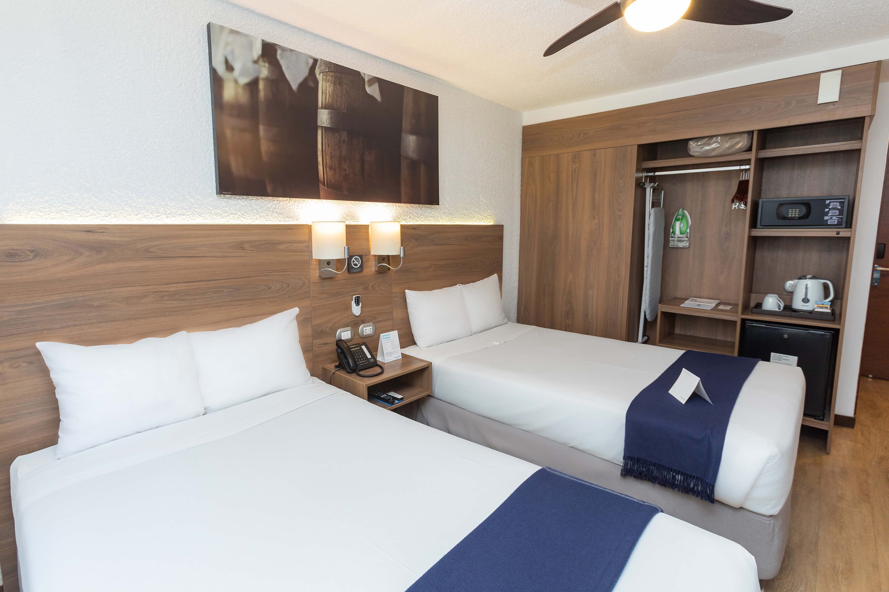 Een hotelkamer met twee bedden, een houten hoofdeinde, een plafondventilator, een magnetron, een minibar en een klein kastje.
