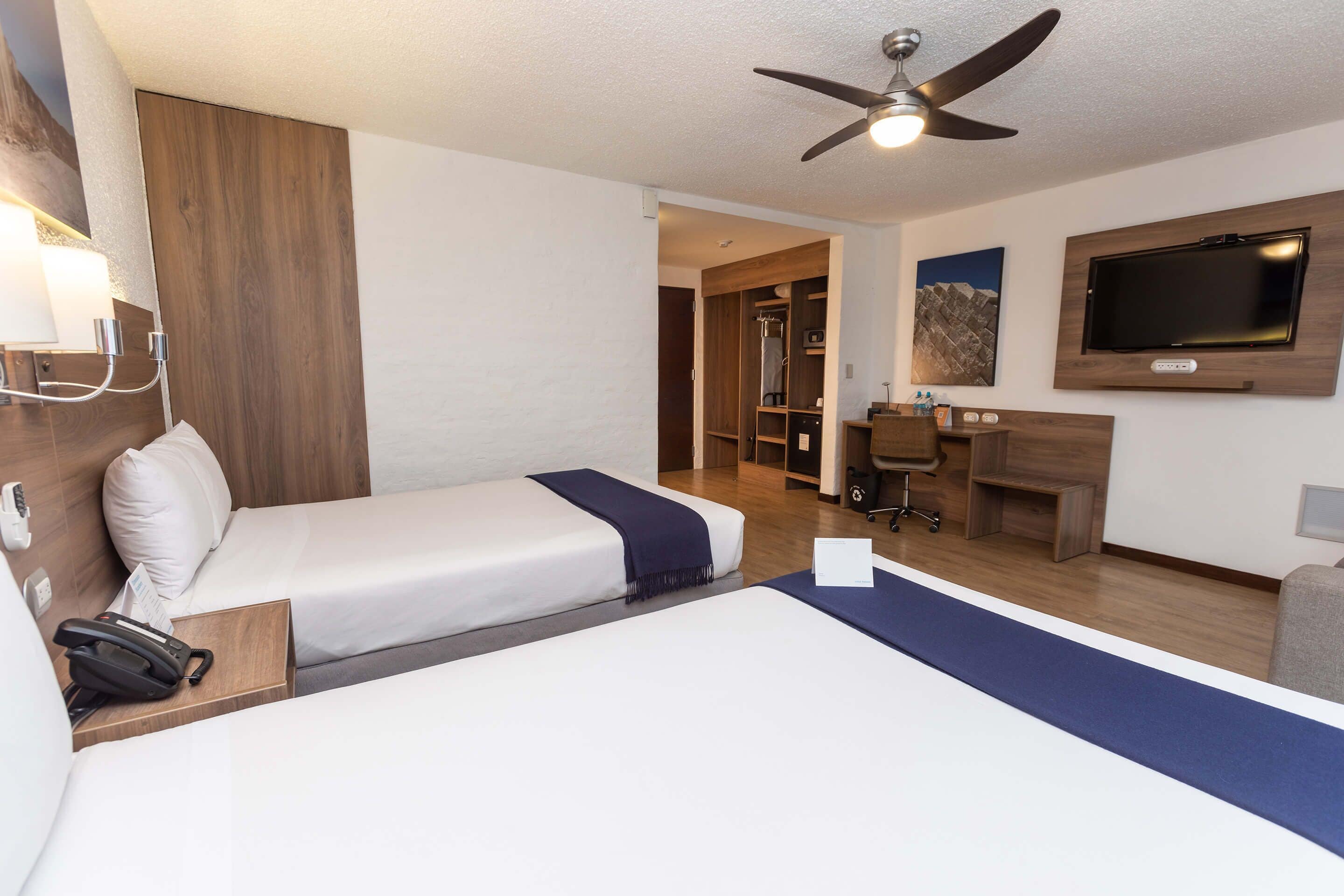 A hotel room with two beds, a desk, a TV, and a ceiling fan.