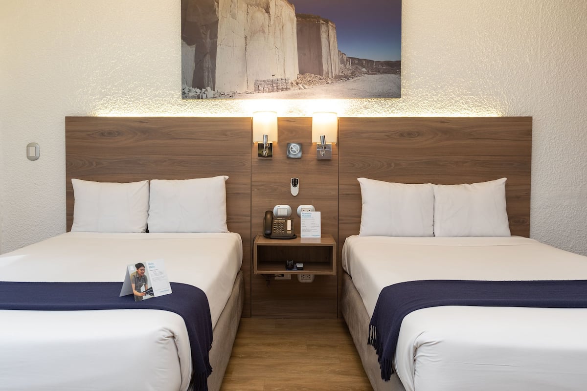 Ein Doppelzimmer mit zwei Betten, einem Holzkopfteil, Nachttischlampen, einem Nachttisch mit Telefon und Speisekarte sowie einem gerahmten Bild an der Wand.
