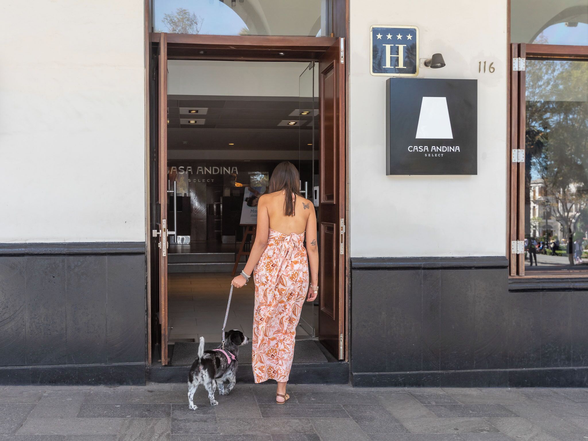 A woman in a floral dress exiting a building with a dog on a leash, and a sign indicating 'CASA ANDINA SELECT'.