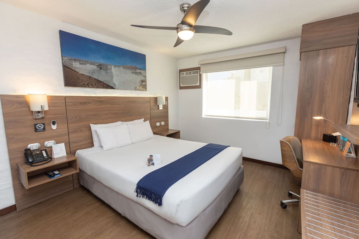 Ein Hotelzimmer mit einem großen Bett, einem Schreibtisch, einem Stuhl, einem Deckenventilator, einem Bild an der Wand und einem Fenster mit Jalousien.