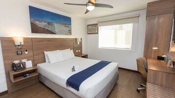 A hotel room with a large bed, a desk, a chair, a ceiling fan, a painting on the wall, and a window with blinds.