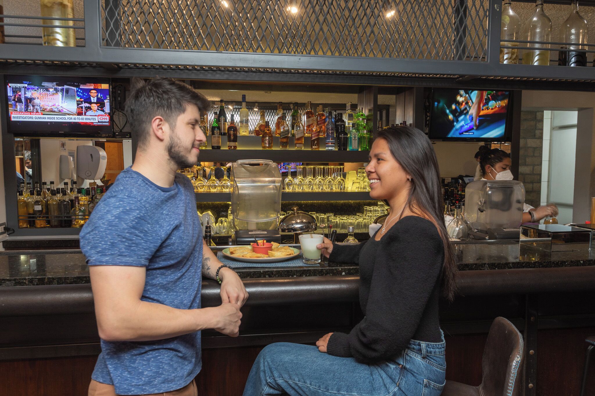 Uma área de bar com balcão, diversas garrafas e uma tela de televisão exibindo notícias.