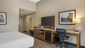 A hotel room with a bed, a desk with a computer, a microwave, and a refrigerator. There are framed pictures on the wall and a lamp on the desk.