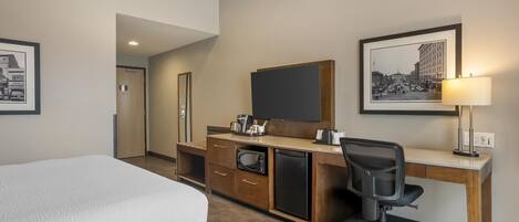 Habitación de hotel con cama, escritorio con computadora, microondas y refrigerador. Hay cuadros en la pared y una lámpara sobre el escritorio.