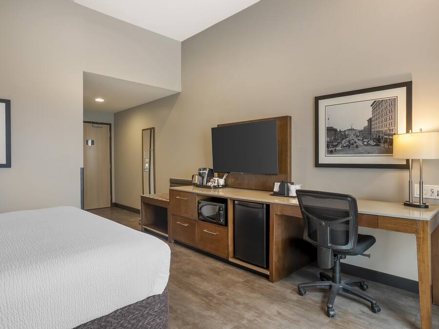 A hotel room with a bed, a desk with a computer, a microwave, and a refrigerator. There are framed pictures on the wall and a lamp on the desk.