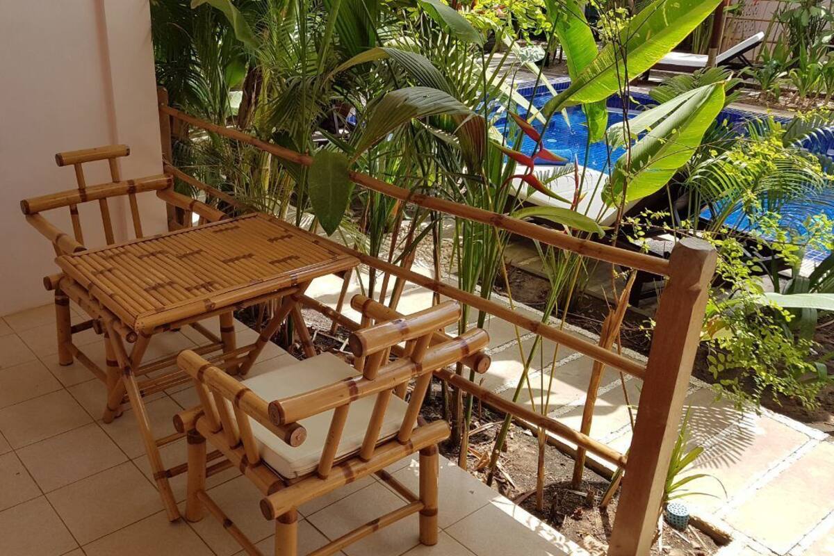 A bamboo furniture set with a table and chairs on a balcony with a view of a pool and tropical plants.