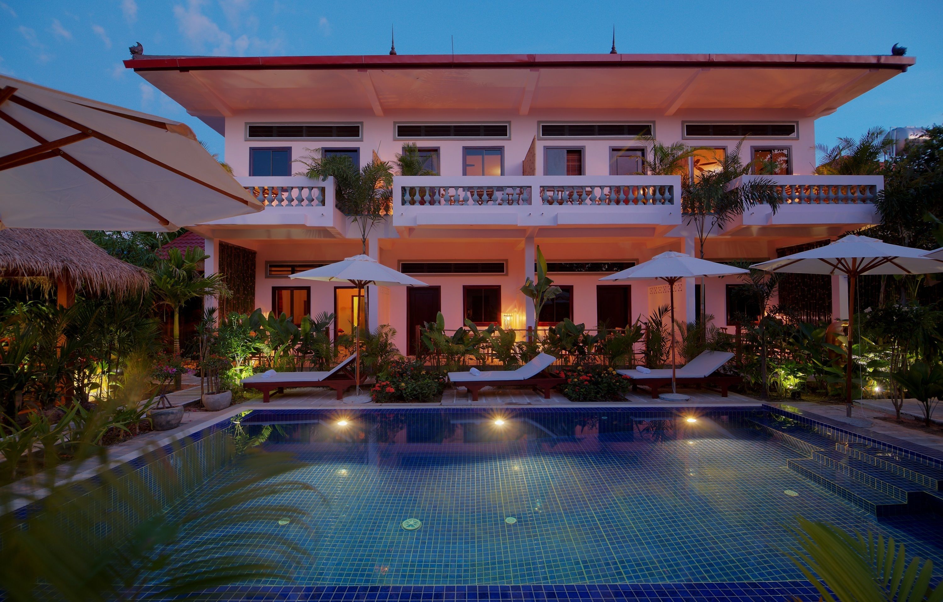 A hotel with a swimming pool, lounge chairs, umbrellas, and a well-lit outdoor area.