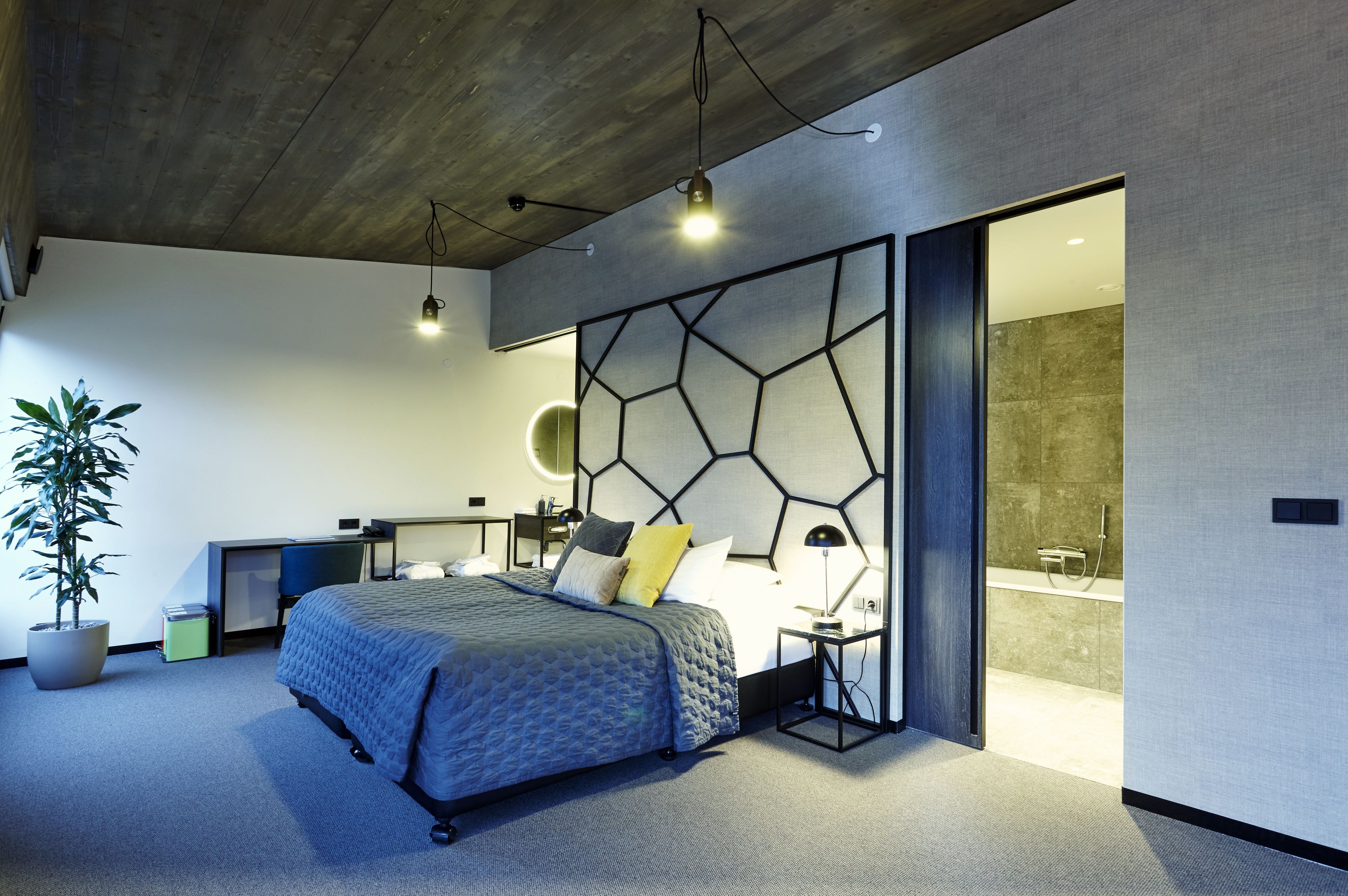 Un dormitorio moderno con una cabecera geométrica, una cama con colcha azul, mesitas de noche, un escritorio y una planta en maceta.