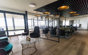 Uma área de estar moderna com diversos tipos de assento, incluindo sofás e poltronas, dispostos em torno de uma mesa de centro. O espaço possui grandes janelas com vista para o exterior, e o teto apresenta um design geométrico único com luminárias pendentes.