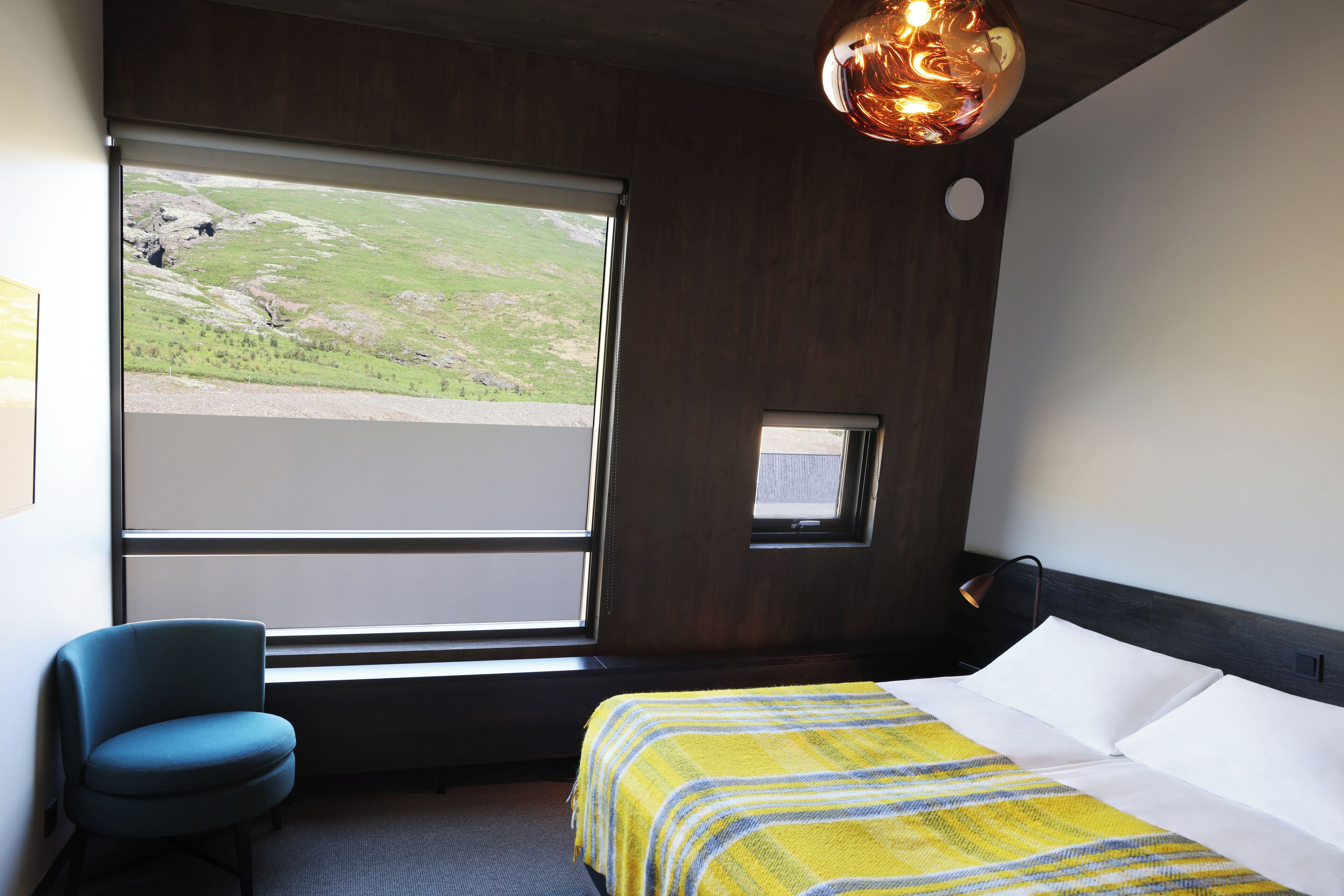 A hotel room with a large window, a bed with a yellow striped blanket, a blue armchair, and a small table lamp.