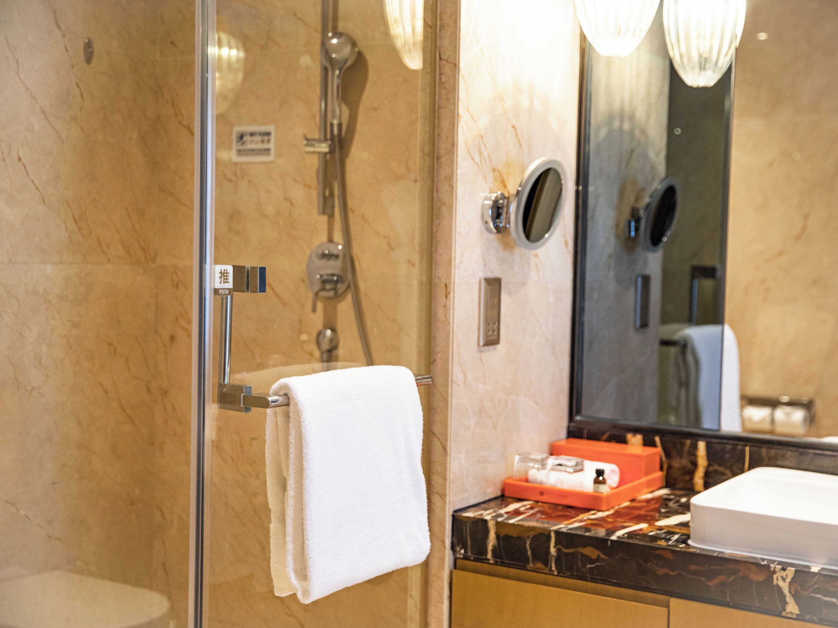 A bathroom with a glass shower enclosure, a white towel hanging on a rack, a mirror, and a marble countertop with toiletries.