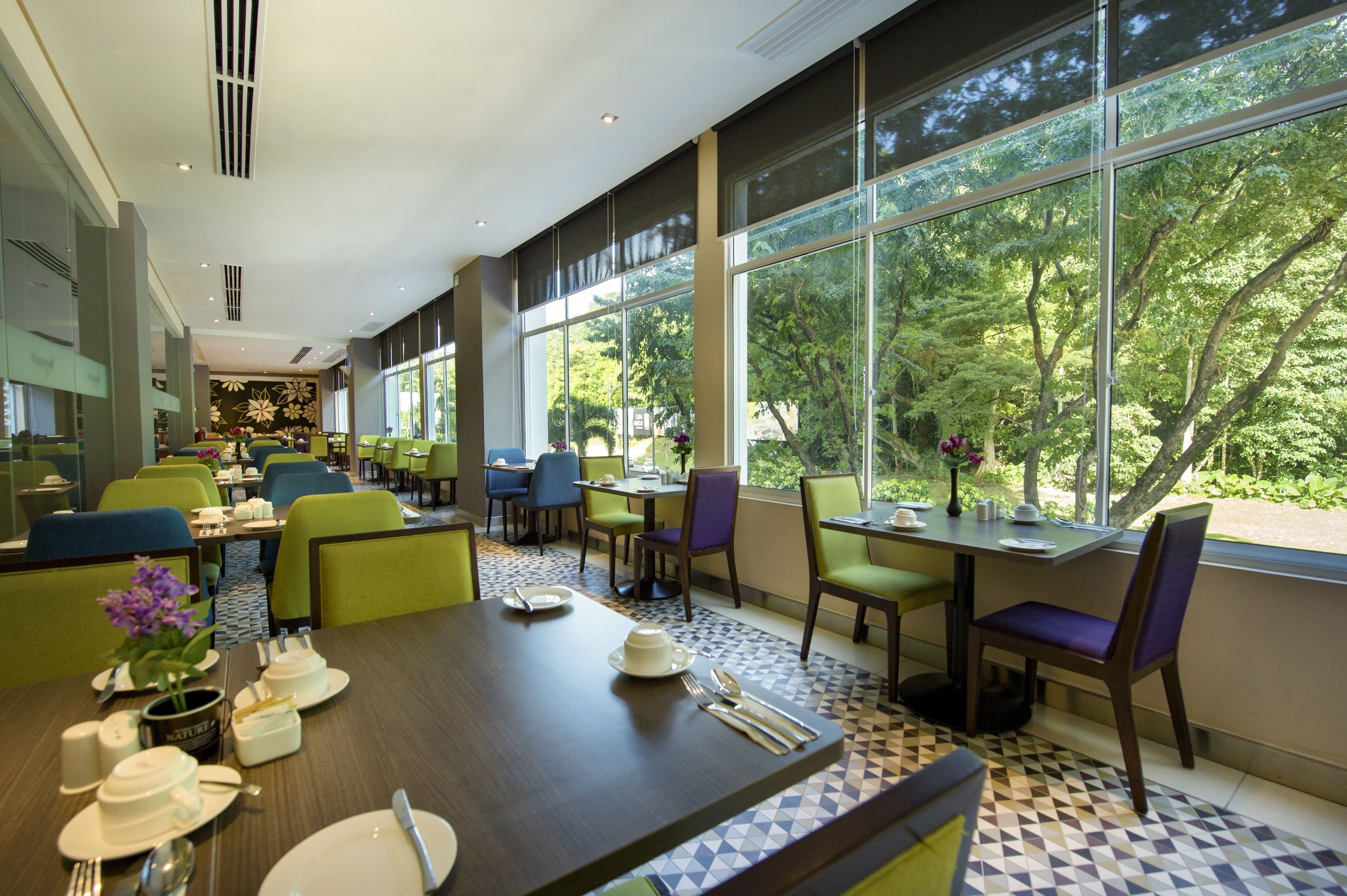 A modern dining area with large windows, green and blue chairs, and neatly set tables.