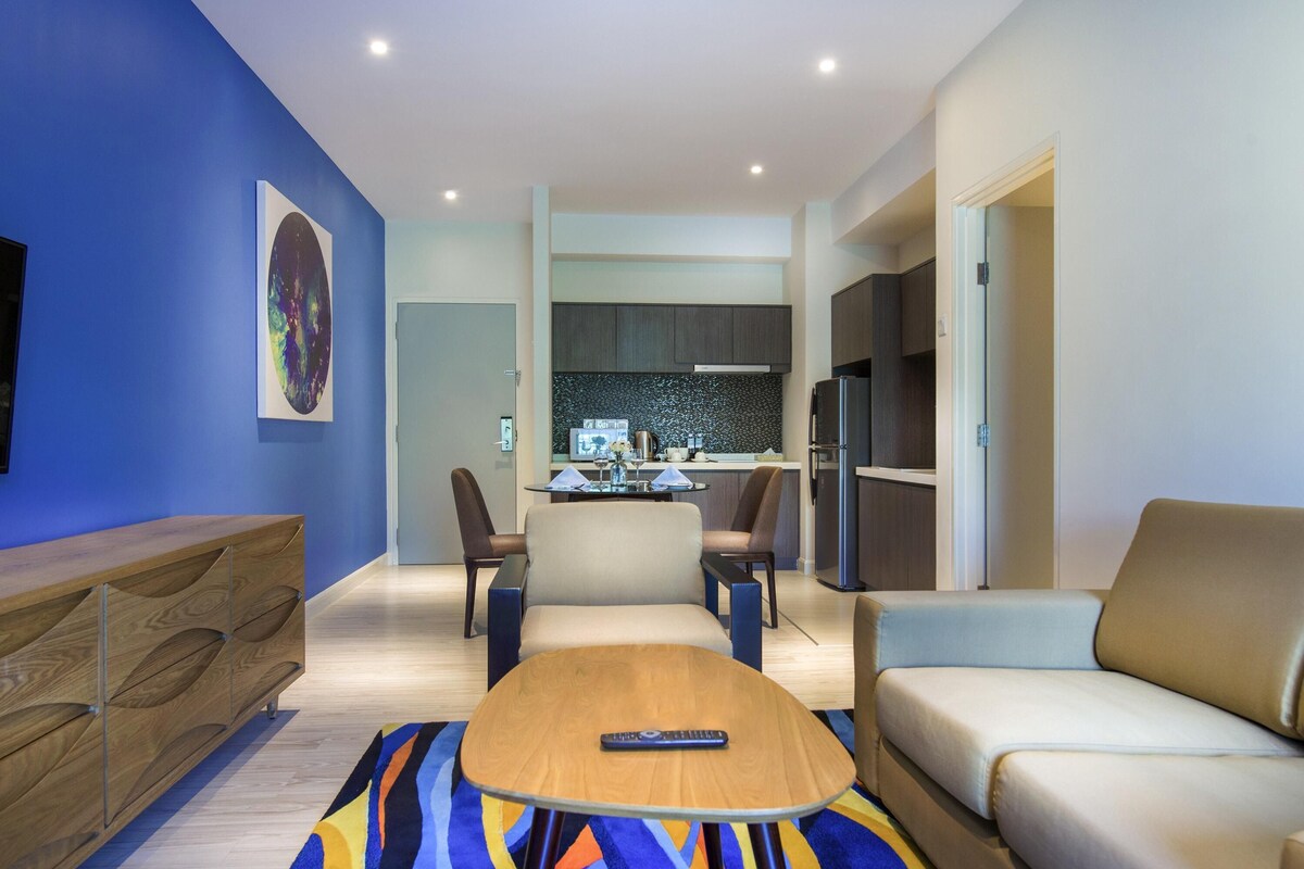A modern living room with a sofa, a coffee table, and a TV. There is a kitchen area in the background with a refrigerator and a microwave.