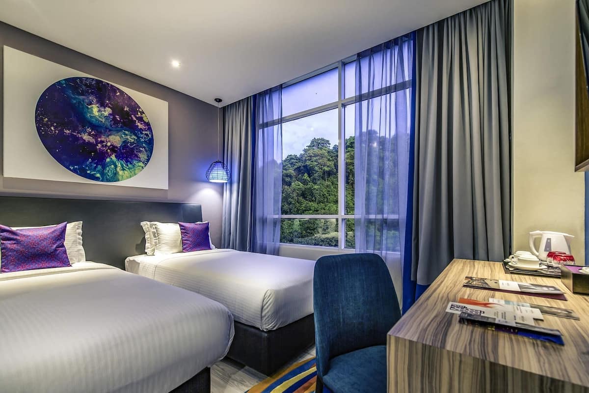 A hotel room with two beds, a desk with a chair, a large window with curtains, and a wall-mounted artwork.