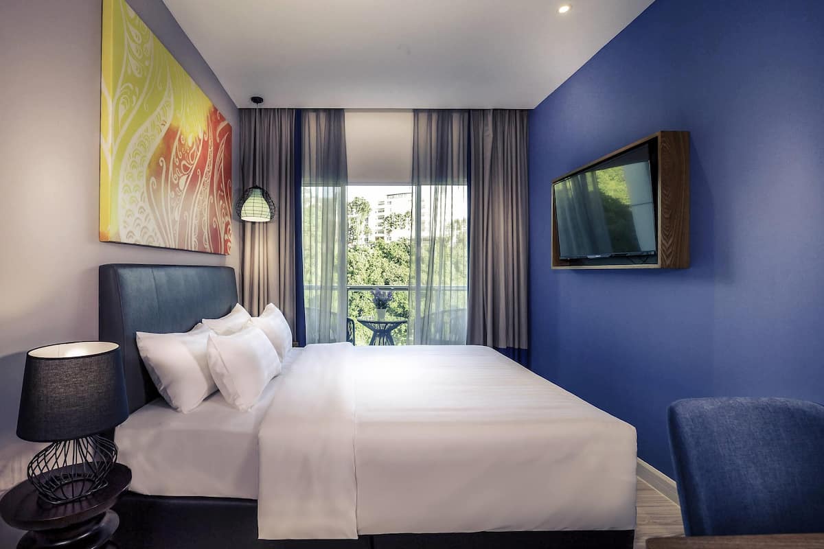 A modern hotel room with a large bed, a desk, a chair, a TV, and a balcony with a view of greenery.