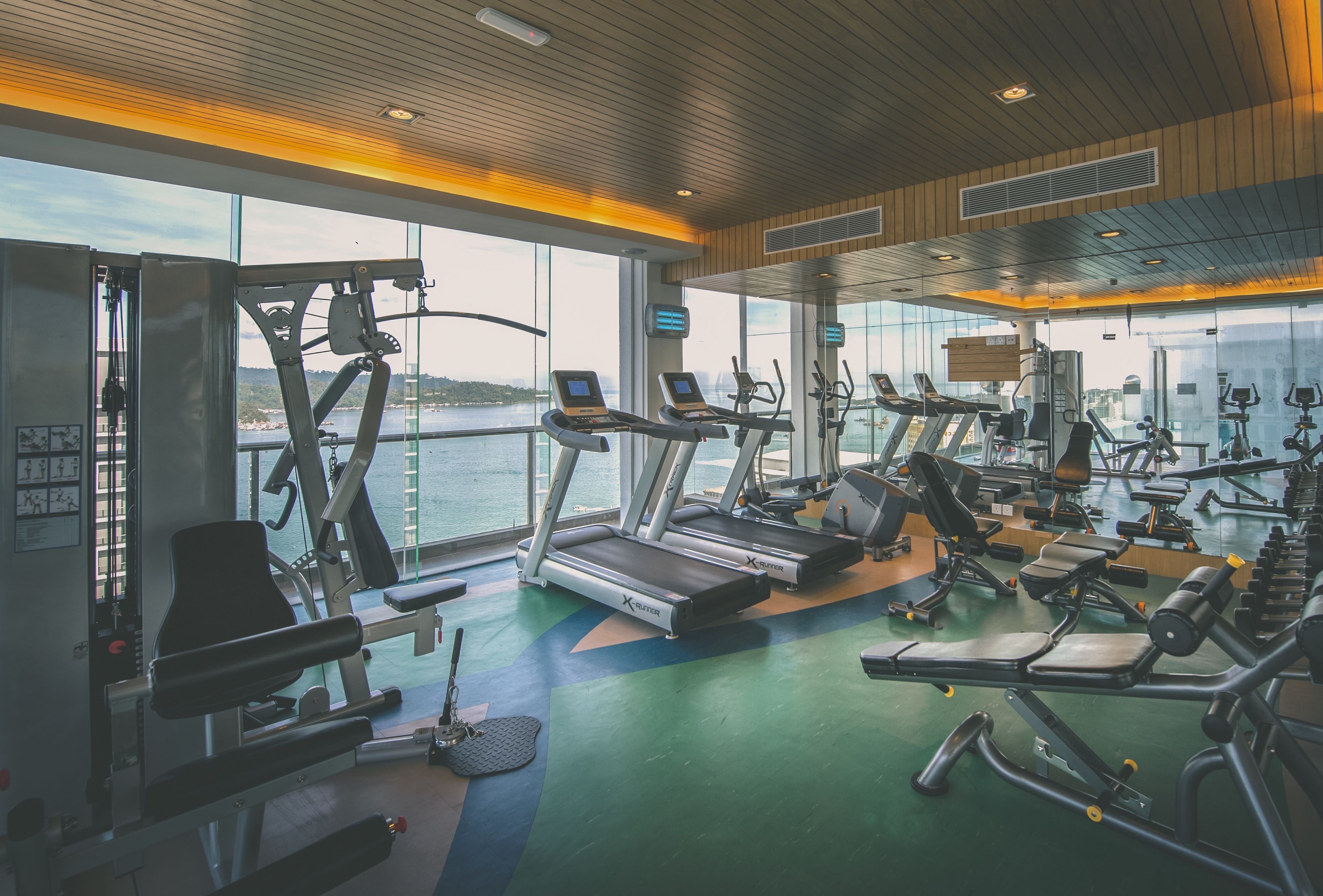 A gym with treadmills, weight machines, and exercise mats, with large windows overlooking a body of water.