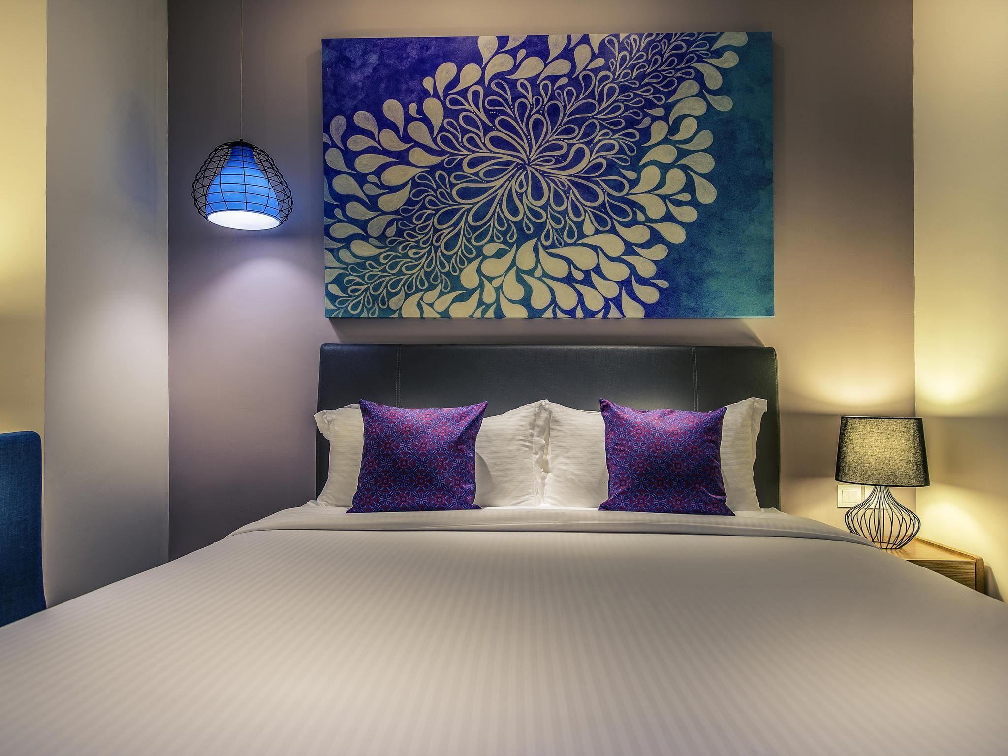 A hotel room with a large bed, a headboard, a wall-mounted artwork, a hanging light fixture, and a bedside table with a lamp.