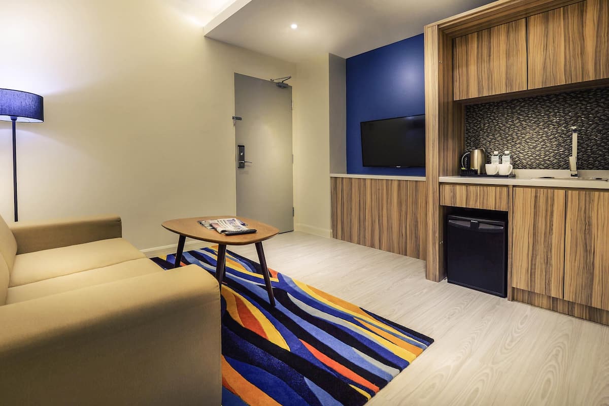 A modern hotel room with a sofa, a small table with a magazine, a TV, and a kitchenette.