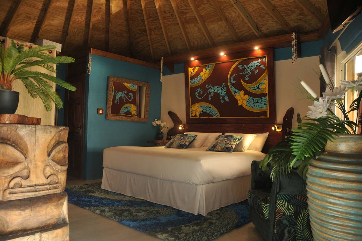 Un dormitorio con techo de madera, una cama con ropa blanca, pared azul y decoración tropical.