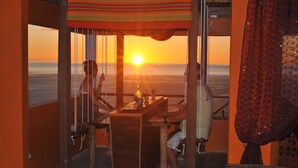 Een kamer met uitzicht op de ondergaande zon boven de oceaan, een houten tafel met stoelen en een kamerkleed met patroon.