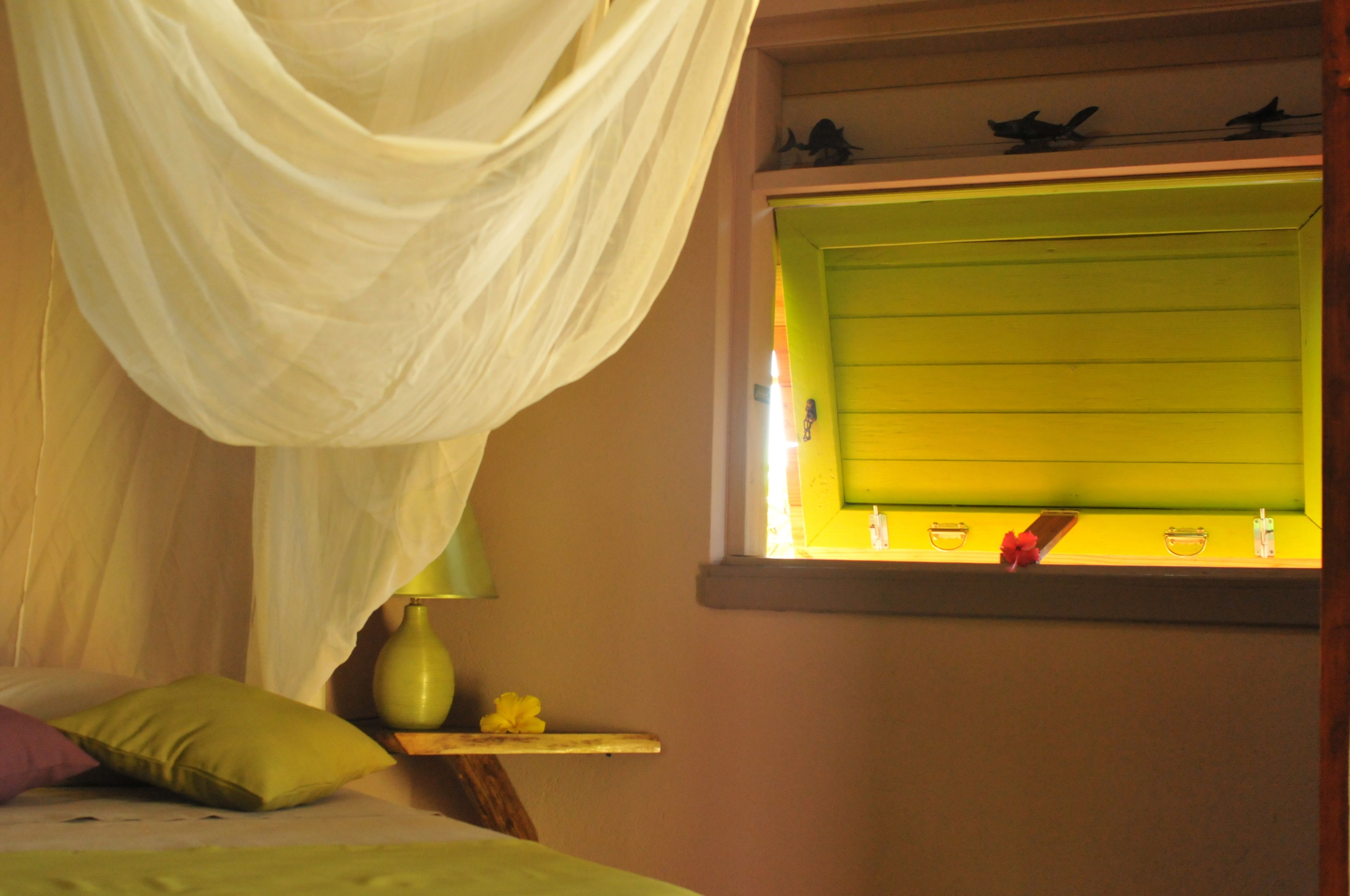 A room with a bed, a window with a green blind, a bedside table with a vase and a flower, and a curtain.