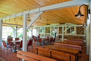 An outdoor covered dining area with wooden benches and tables, stone walls, and a wooden ceiling with hanging lights. - Kuźnia Pałacowa (Korczew)