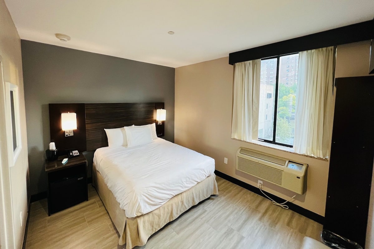 A hotel room with a large bed, bedside lamps, a nightstand, a window with curtains, and a wall-mounted heater.