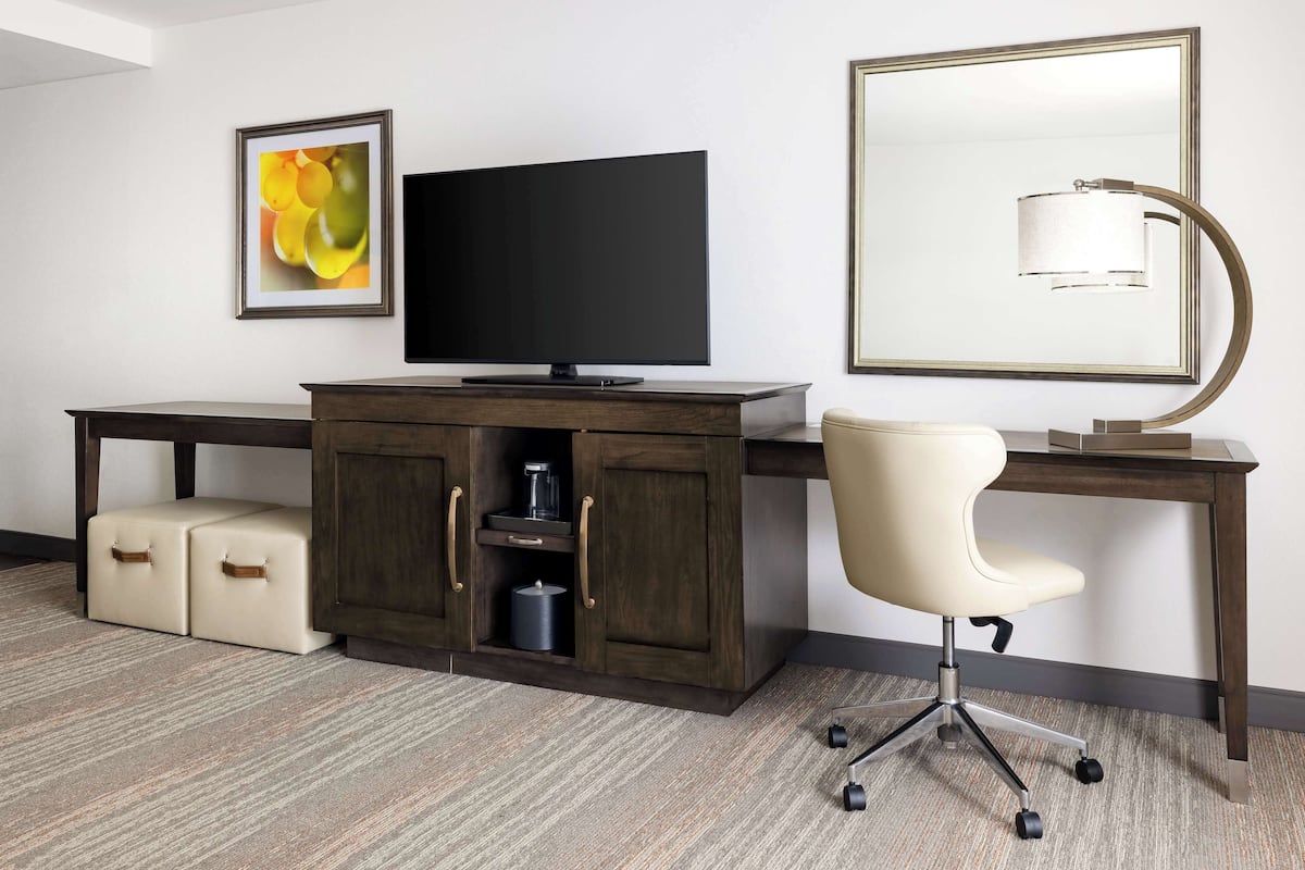 A hotel room with a dark wooden desk, a flat-screen TV, a white office chair, a framed picture on the wall, and a floor lamp.