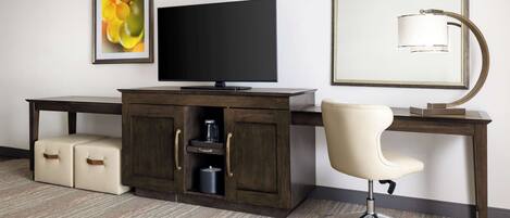 A hotel room with a dark wooden desk, a flat-screen TV, a white office chair, a framed picture on the wall, and a floor lamp.